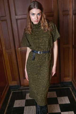 A woman wearing a short-sleeved olive green patterned dress with a black belt, standing in front of dark wood paneling and a checkered floor.