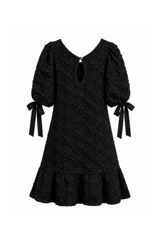 Back of black embroidered babydoll dress with keyhole cut-out on neckline, puff sleeves, bows at cuffs and ruffle hem, shown against white background. 