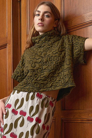A woman leans against a wooden paneled wall wearing a cropped olive green quilted knit top and a cream skirt with a red and olive green radish pattern.
