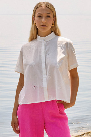 Woman in front of lake wearing white mandarin collar button up blouse with pink pants. 