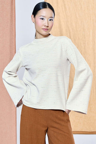 A woman modeling a cream-colored, textured knit mock-neck top with wide bell sleeves, paired with brown trousers, set against a split beige and light wood-paneled background.