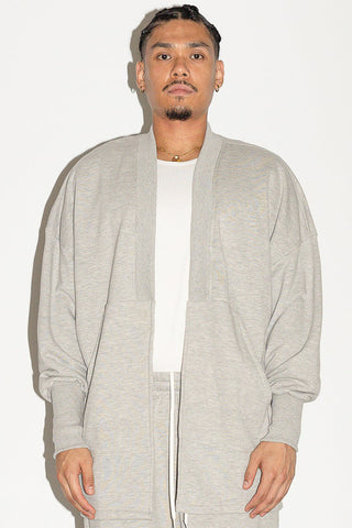 Man in front of white background wearing grey open front cardigan over white shirt. 