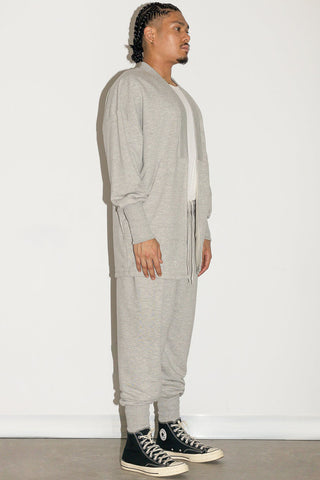 Man in front of white background wearing grey open front cardigan over white shirt with matching grey track pants. 