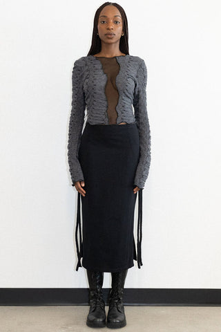 Woman in front of white background wearing fitted black midi skirt with grey textured long sleeve Hallow top by Mercy House, featuring a mesh insert. 