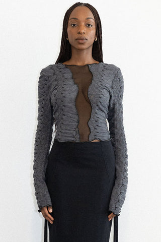 Woman in front of white background wearing fitted black skirt with grey textured long sleeve Hallow top by Mercy House, featuring a mesh insert. 