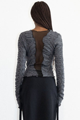 Side view of woman in front of white background wearing fitted black skirt with grey textured long sleeve Hallow top by Mercy House, featuring a mesh insert. 