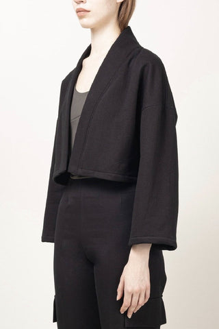 A model wearing a black cropped jacket with wide three-quarter sleeves, styled over a dark gray top and black trousers, photographed against a plain off-white studio background.
