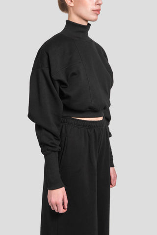 A model shown from the side wearing a black cropped mock-neck sweatshirt and matching black wide-leg sweatpants against a solid light gray background.