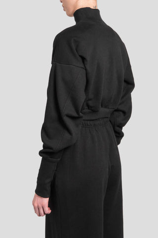 A model shown from the back wearing a black mock-neck cropped sweatshirt and matching black wide-leg trousers against a solid light gray background.