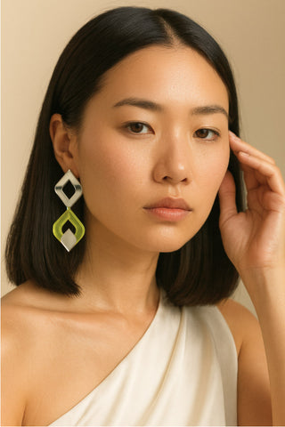 A woman with dark shoulder-length hair wearing a white one-shoulder top and a large geometric dangling earring with white and lime green shape. 