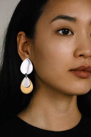 A close-up of a woman wearing a pair of teardrop-shaped dangle earrings made from silver and gold plexiglass. 