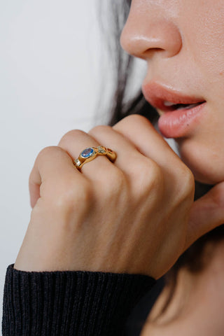 Close-up of woman's face and hand with gold ring and multiple colourful stones. 