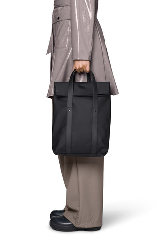 A person wearing a shiny taupe trench coat and taupe trousers holds a black top-handle tote bag against a clean white background.
