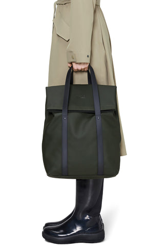 A person wearing a beige trench coat and glossy black rain boots holds a dark olive green tote bag with black handles against a clean white background.