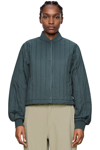 Woman wearing a dark teal-green cropped quilted jacket with a high collar and beige trousers, shot against a plain white background.