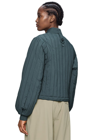 A woman shown from the back wearing a dark teal quilted jacket with vertical stitching and beige trousers, set against a plain white background.