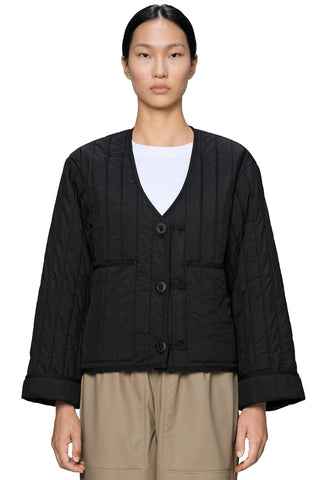A woman wearing a black quilted liner jacket, styled over a white t-shirt and beige trousers, against a plain white background.