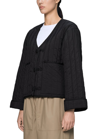 A woman wearing a black quilted cropped liner jacket with v-neck and button closure, styled with a white t-shirt and beige trousers. 