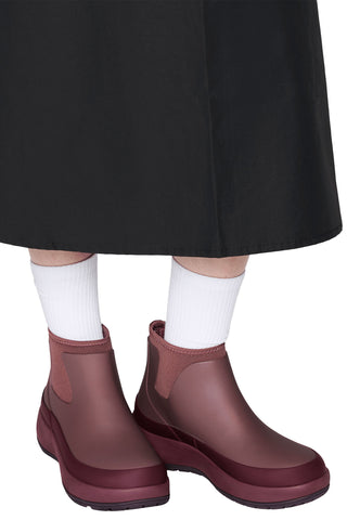 A close-up of a person's feet wearing a pair of dark reddish-brown rubber rain boots with white crew socks and a black skirt, against a plain white background.
