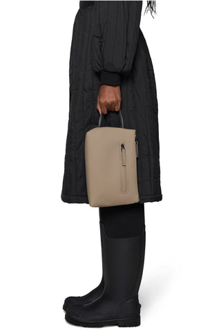 Person wearing black coat holding dark beige cosmetic bag with black handle. 