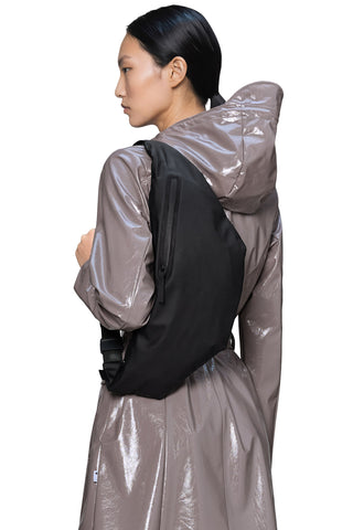 Back view of woman wearing a glossy taupe-brown hooded raincoat and a black oversized bum bag, draped over her shoulder. 