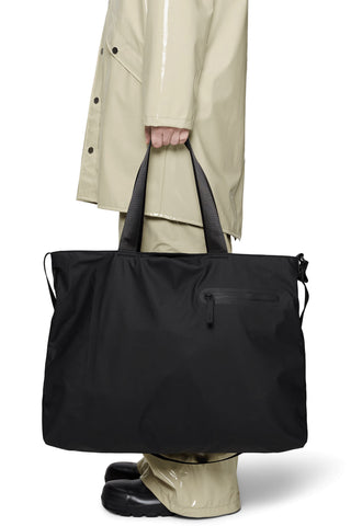 Person wearing beige coat holding black messenger tote bag with carry handles. 