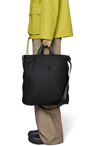 A person wearing a bright yellow-green jacket and beige trousers holds a large black nylon tote bag with top handles and a shoulder strap against. 