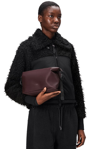 Woman in front of white background wearing black zip up sweater and burgundy toiletry bag. 