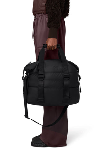Close-up of woman in front of white background wearing burgundy pants, holding black quilted weekend bag. 