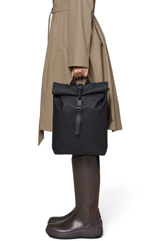A person wearing a beige trench coat and dark brown rubber rain boots holds a black waterproof roll-top backpack against a plain white background.