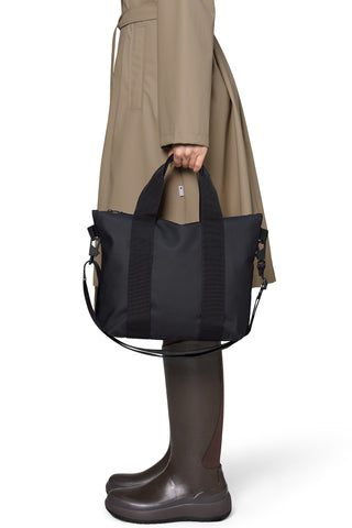 A person wearing a beige trench coat and dark brown rain boots holds a small black tote bag with double top handles and a shoulder strap against a white studio background.