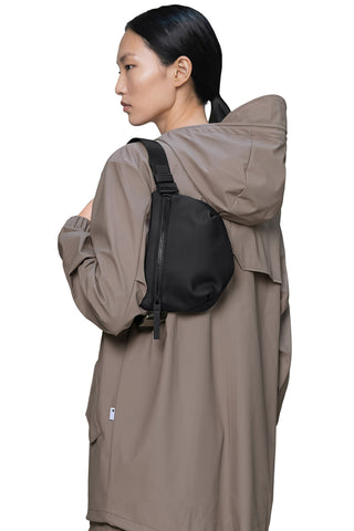 A woman wearing a taupe-brown hooded jacket and a black crossbody sling bag, shown from a side-back angle against a solid white background.