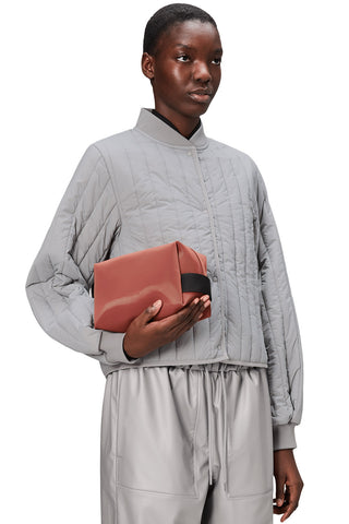 Woman in front of white background wearing grey quilted jacket, grey pants and holding clay pink coloured toiletry bag. 