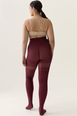 Back view of woman in front of white background wearing beige bra and burgundy over-the-knee tights. 