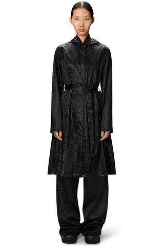 Woman, standing in front of white background, wearing shiny black Aline belted Longer Jacket by RAINS. 