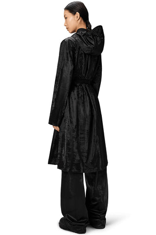 Back view of woman, standing in front of white background, wearing shiny black Aline belted Longer Jacket by RAINS. 