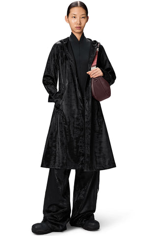 Woman, standing in front of white background, wearing unzipped shiny black Aline belted Longer Jacket by RAINS with dark red bag. 