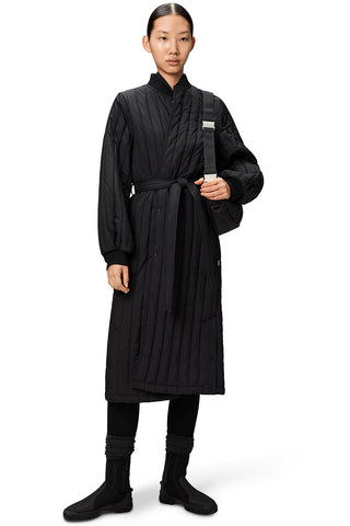 Woman, in front of white background, wearing black quilted and belted long Banja Liner Coat by RAINS. 