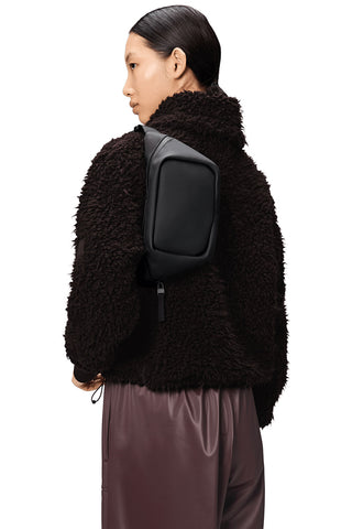 Woman in front of white background, back to camera, wearing a fuzzy black jacket and Black Bum Bag Mini by RAINS. 