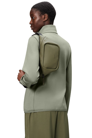 Woman in front of white background, back to camera, wearing light green jacket and Marsh green Bum Bag Mini by RAINS. 