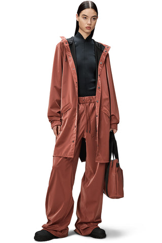 Woman wearing clay coloured unzipped unisex waterproof RAINS long jacket with matching pants and bag, against white background. 