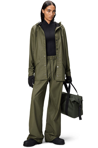 Woman standing in front of white background wearing unzipped Marsh (green) unisex waterproof RAINS jacket with matching pants and bag. 