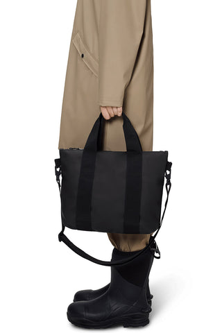 Waist down side view image in front of white background of a woman wearing long beige coat and holding a RAINS Tote Bag Micro in black. 