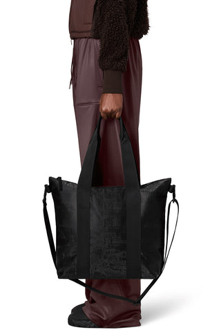 Waist down side view image in front of white background of a woman wearing all burgundy and holding a RAINS Tote Bag Micro in Coal black colour. 