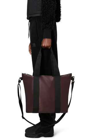 Waist down side view image in front of white background of a woman wearing all black and holding a RAINS Tote Bag Micro in deep red Depth colour. 