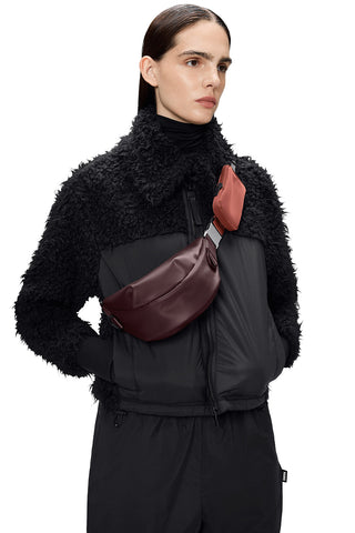 Woman wearing fluffy black zip up acket with deep red Depth Valera Shoulder Bag Mini by RAINS. 