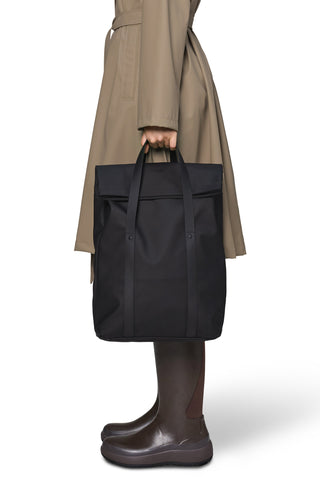 A person wearing a beige trench coat and dark brown rain boots holds a large black tote backpack with top handles against a clean white background.