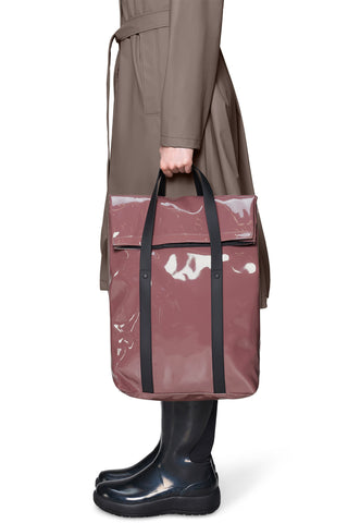 A person wearing a beige trench coat and dark brown rain boots holds a large iridescent pink tote backpack with top handles against a clean white background.