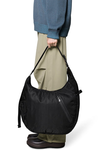 A person wearing a blue sweater, beige pants, and brown boots holds a black nylon shoulder bag with a zippered pocket against a white background.