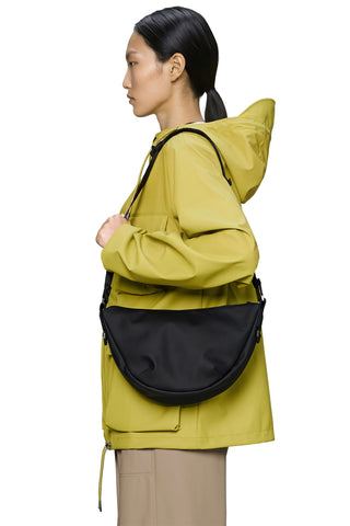 A model in profile wearing a bright yellow-green hooded jacket and beige pants, carrying a black crescent-shaped shoulder bag against a plain white background.
